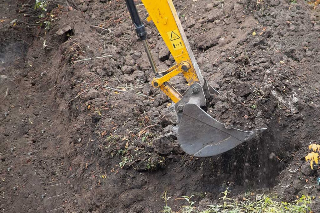 A worker operates an excavator to dig a trench through grassy soil on a rural property.