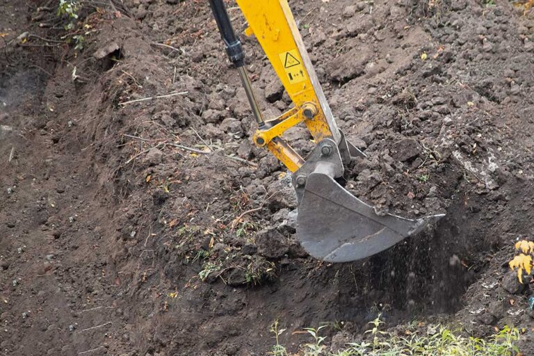 A worker operates an excavator to dig a trench through grassy soil on a rural property.